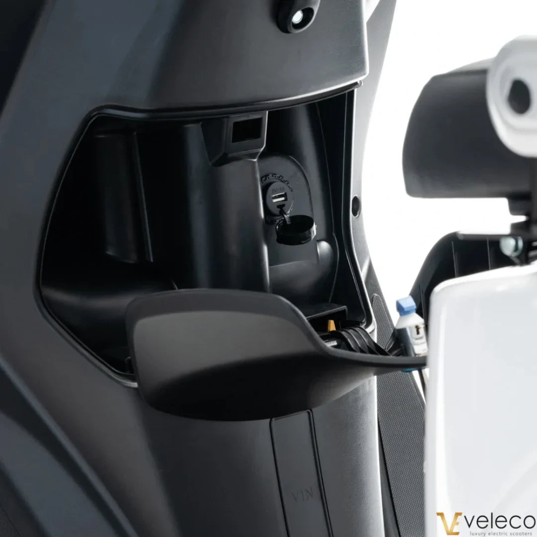 Izdelek na sliki je električni skuter znamke Veleco, model Cristal. Na sliki je prikazan del notranjosti skuterja, kjer so vidni priključki in delovni mehanizmi, kar kaže na sodobno zasnovo in funkcionalnost. Oprema vključuje tudi prostor za polnjenje, kar je pomembno za delovanje električnega vozila.