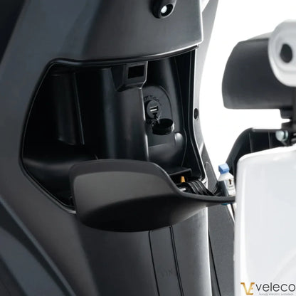 Izdelek na sliki je električni skuter znamke Veleco, model Cristal. Na sliki je prikazan del notranjosti skuterja, kjer so vidni priključki in delovni mehanizmi, kar kaže na sodobno zasnovo in funkcionalnost. Oprema vključuje tudi prostor za polnjenje, kar je pomembno za delovanje električnega vozila.