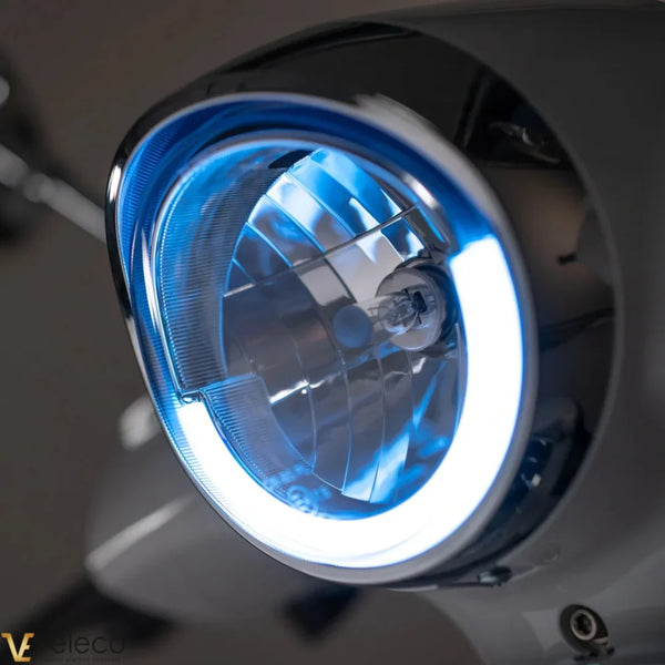 Izdelek na sliki je električni skuter, znamke Veleco, model Cristal. Na sliki je prikazana sprednja luč skuterja, ki ima modro osvetlitev in sodoben dizajn. Luč je zasnovana za izboljšanje vidnosti in varnosti med vožnjo.