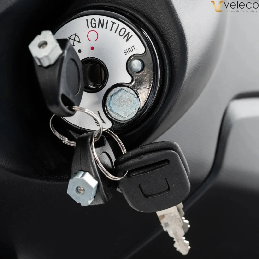 Izdelek je električni skuter, znamke Veleco, model Cristal. Na sliki je prikazan ključavnica za vžig, ki vključuje ključe in oznake za delovanje, kot so "SHUT" in "IGNITION". Vidna je tudi konstrukcija okoli ključavnice, kar nakazuje na funkcionalnost in zasnovo skuterja.