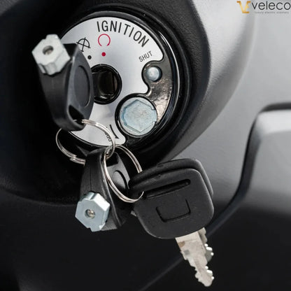 Izdelek je električni skuter, znamke Veleco, model Cristal. Na sliki je prikazan ključavnica za vžig, ki vključuje ključe in oznake za delovanje, kot so "SHUT" in "IGNITION". Vidna je tudi konstrukcija okoli ključavnice, kar nakazuje na funkcionalnost in zasnovo skuterja.