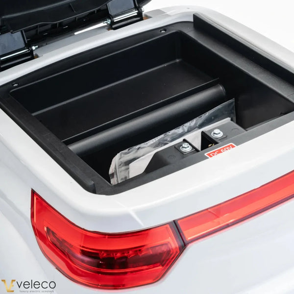 Izdelek je električni skuter, znamke Veleco, model Cristal. Na sliki je prikazan odprt prostor za shranjevanje, ki razkriva baterijo in notranjost skuterja. Vidna je tudi zadnja luč skuterja, kar nakazuje na njegov dizajn in funkcionalnost.