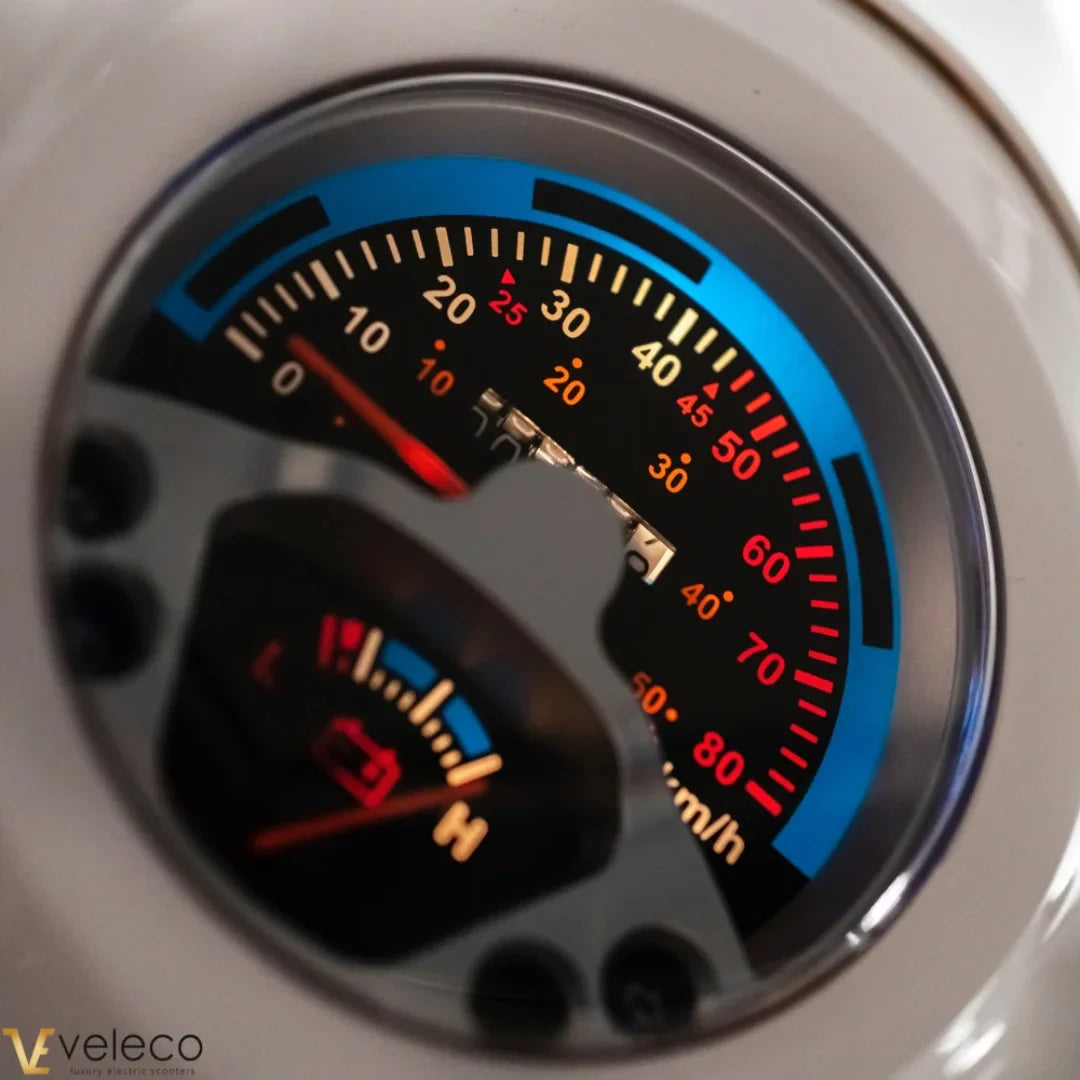 Izdelek na sliki je električni skuter znamke Veleco, model Cristal. Na sliki je prikazan merilnik hitrosti, ki prikazuje različne vrednosti hitrosti in nivoja baterije. Merilnik ima barvne oznake, ki omogočajo enostavno branje podatkov med vožnjo.