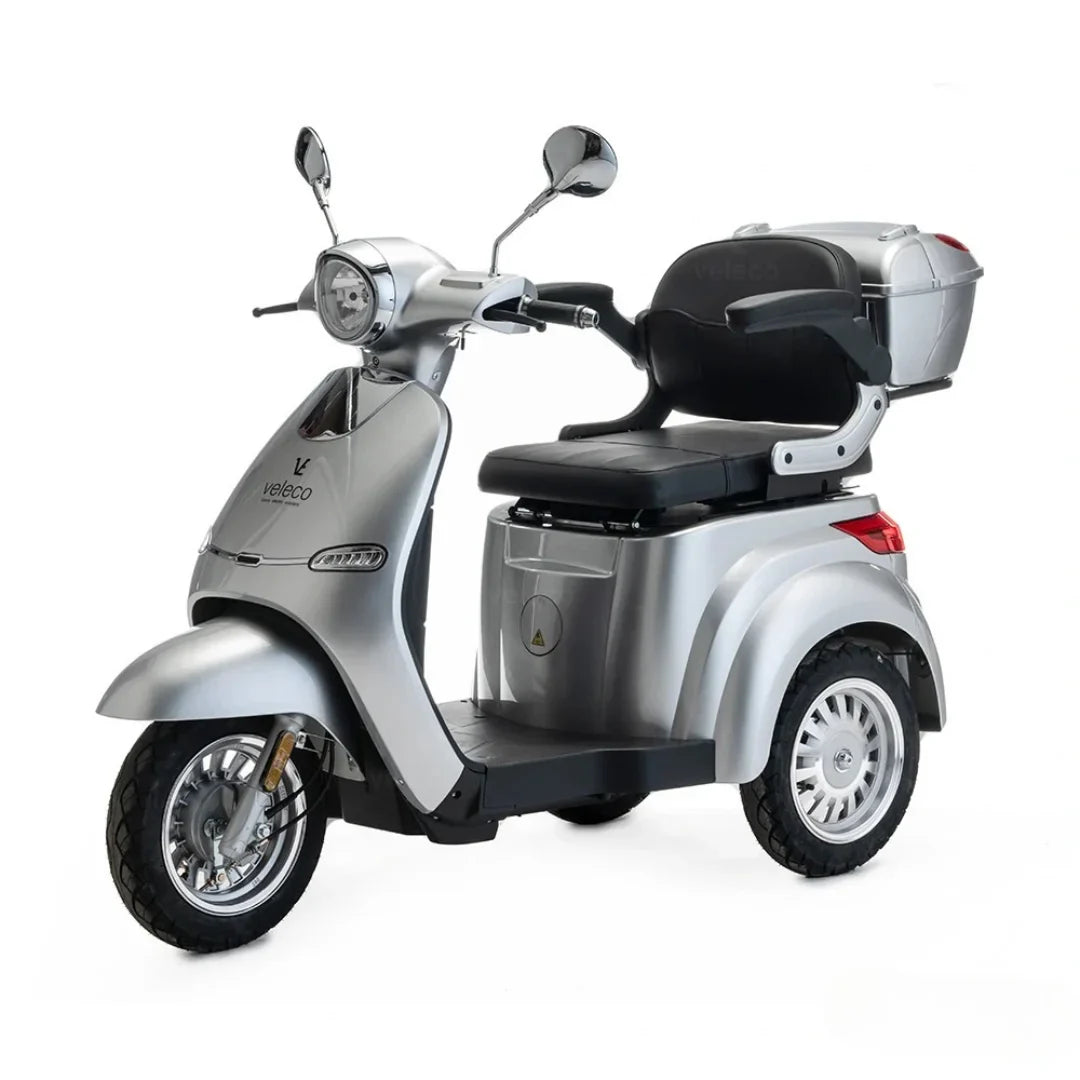 Izdelek na sliki je električni mobilni voziček znamke Veleco, model Cristal. Gre za električni skuter s tri kolesi, ki je zasnovan za udobno in varno vožnjo. Na sliki je vidna srebrna karoserija, udobno sedežno mesto ter prtljažnik na zadnjem delu vozila, kar omogoča enostavno shranjevanje osebnih stvari.