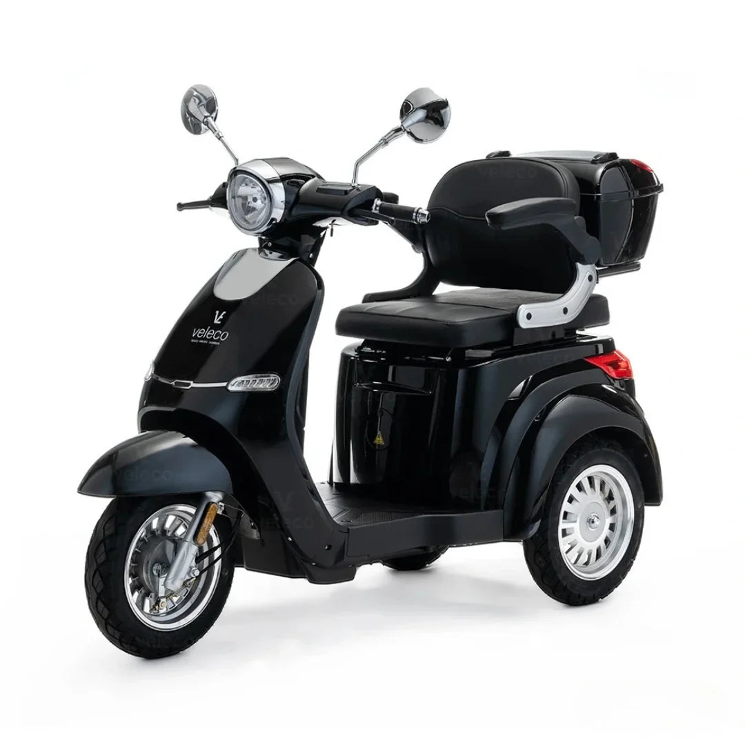 Izdelek je električni skuter, model Veleco Cristal. Na sliki je vidna črna električna vozila s tremi kolesi, opremljena z udobnim sedežem in prtljažnikom na zadnjem delu. Skuter ima klasičen dizajn z retro elementi, vključno s kromiranimi ogledali in sprednjim svetlom.
