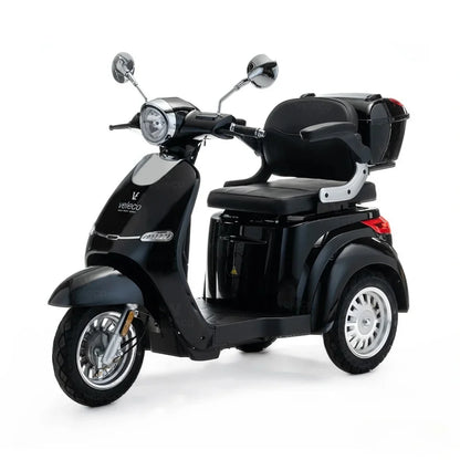 Izdelek je električni skuter, model Veleco Cristal. Na sliki je vidna črna električna vozila s tremi kolesi, opremljena z udobnim sedežem in prtljažnikom na zadnjem delu. Skuter ima klasičen dizajn z retro elementi, vključno s kromiranimi ogledali in sprednjim svetlom.