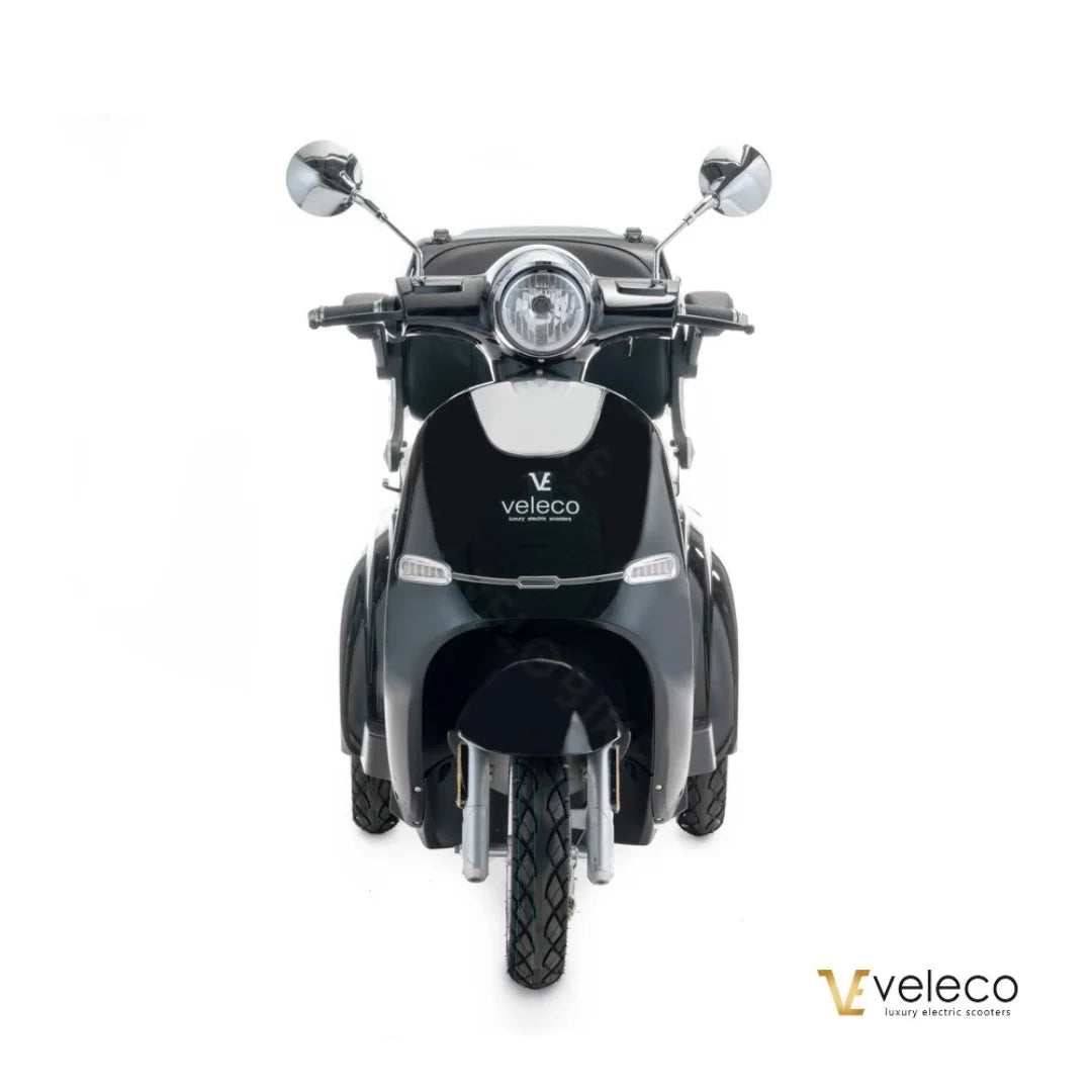 Izdelek je električni skuter znamke Veleco, model Cristal. Na sliki je prikazan sprednji del skuterja, ki ima retro obliko z značilnimi kromiranimi ogledali in okroglo sprednjo lučjo. Barva skuterja je temna, kar mu daje eleganten videz. Na zadnjem delu je jasno vidna oznaka znamke Veleco.