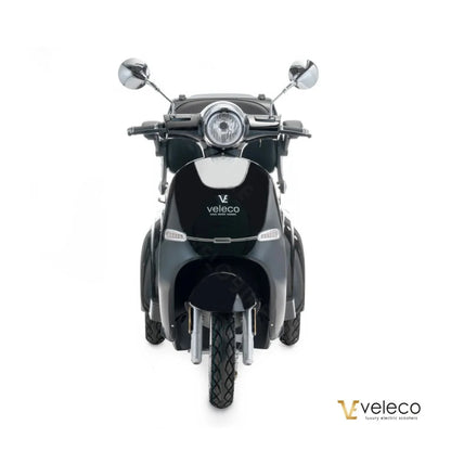 Izdelek je električni skuter znamke Veleco, model Cristal. Na sliki je prikazan sprednji del skuterja, ki ima retro obliko z značilnimi kromiranimi ogledali in okroglo sprednjo lučjo. Barva skuterja je temna, kar mu daje eleganten videz. Na zadnjem delu je jasno vidna oznaka znamke Veleco.