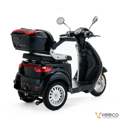 Izdelek na sliki je električni skuter, model Veleco Cristal. Skuter je črne barve in ima prostoren zadnji prtljažnik, kar omogoča udobno shranjevanje. Na sliki so vidni tudi ogledali in nadzorna plošča, ki vključuje ključavnico. Skuter je zasnovan za udobno vožnjo in enostavno upravljanje.