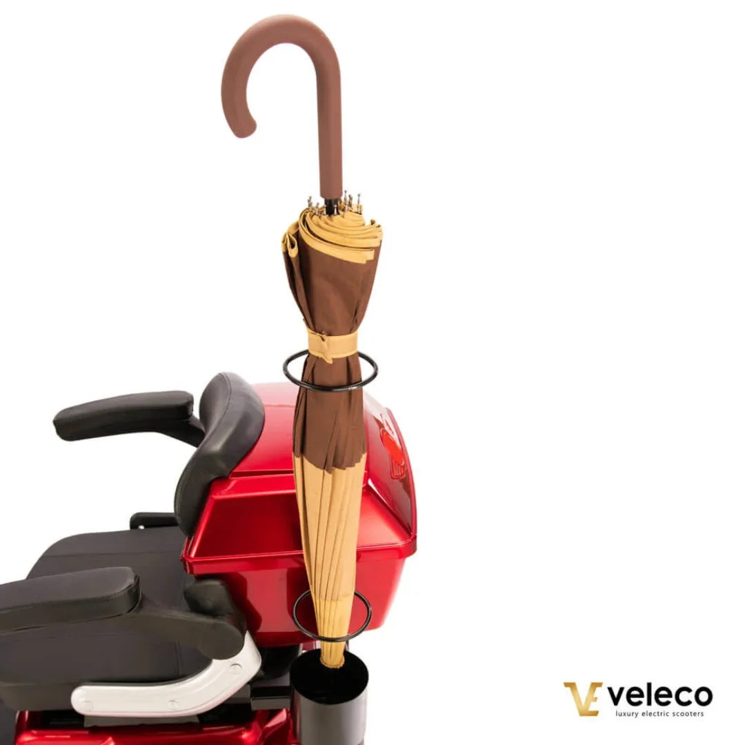 Izdelek na sliki je električni skuter, model Veleco Draco. Na sliki je vidna rdeča barva skuterja, ki ima udobno sedežno površino in naslonjala za roke. Na zadnjem delu skuterja je pritrjen dežnik, kar nakazuje na praktičnost in udobje pri uporabi v različnih vremenskih razmerah.