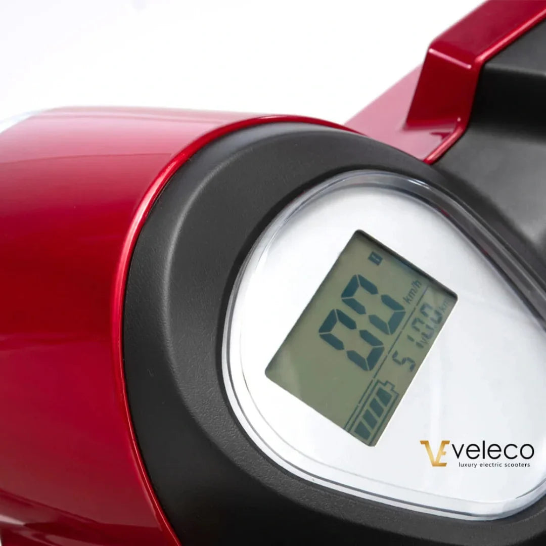 Izdelek na sliki je električni skiro, znamke Veleco, model Draco. Na sliki je prikazan del armaturne plošče, ki vključuje digitalni zaslon s prikazom hitrosti, ki trenutno znaša 0 km/h. Barva skiroja je rdeča, kar mu daje privlačen videz.