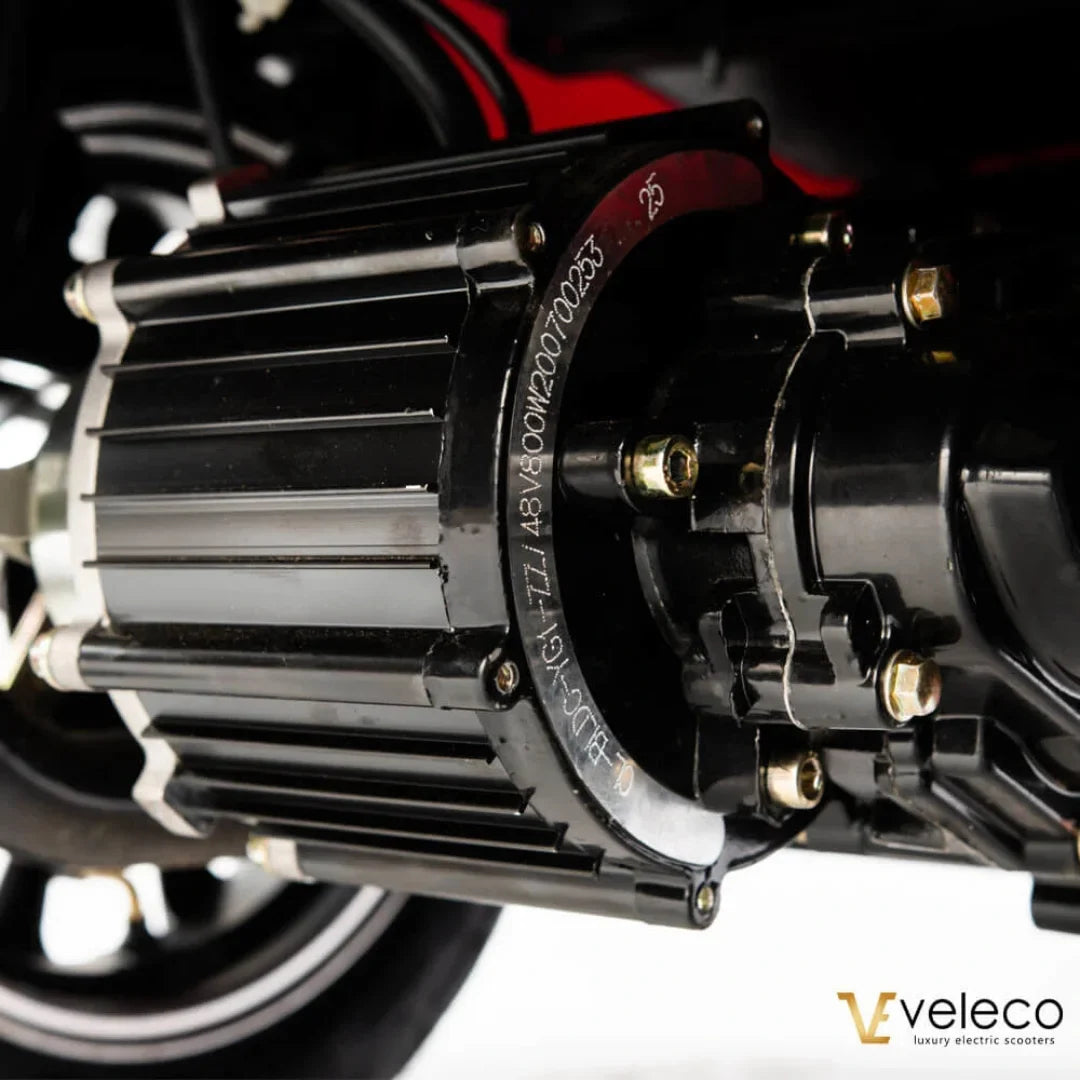 Izdelek je električni skuter, model Veleco Draco. Na sliki je prikazan motor skuterja, ki je črn in ima značilno obliko z rešetkami za hlajenje. Vidna sta tudi del kolesa in nekateri kabli, kar nakazuje na tehnično zasnovo in funkcionalnost vozila.