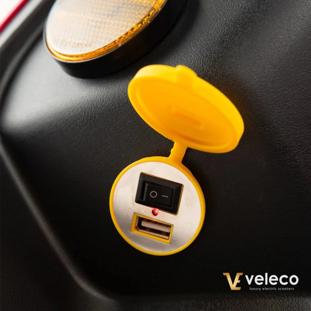 Izdelek je električni skiro, model Veleco Draco. Na sliki je prikazan del skiroja, kjer je vidna rumena zaščitna kapica, ki pokriva USB priključek in stikalo. Rdeča lučka ob stikalu kaže, da je naprava aktivna. V ozadju je tudi odsevna oznaka.