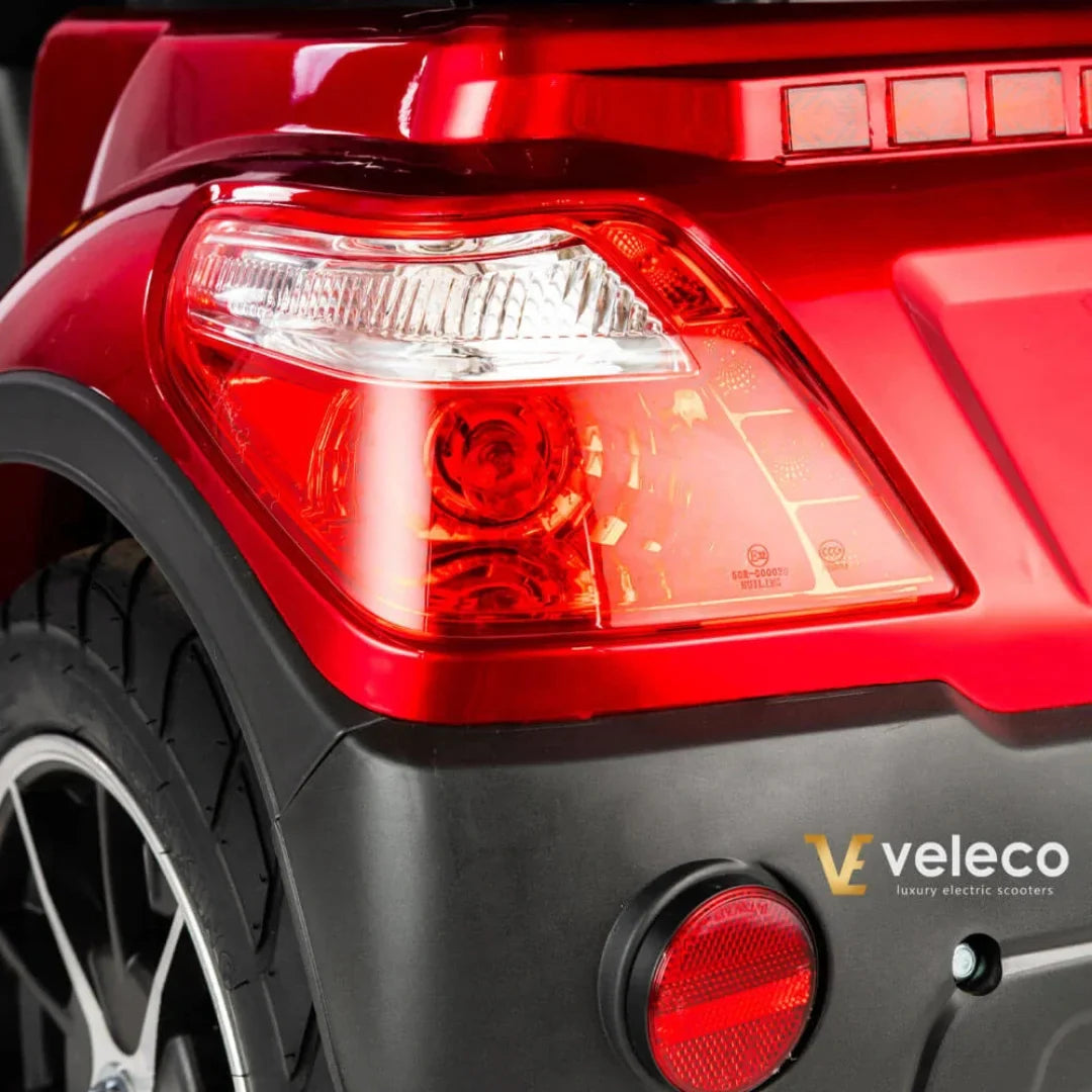 Izdelek je električni skuter, model Veleco Draco. Na sliki je vidna zadnja luč skuterja, ki ima rdečo barvo in je oblikovana tako, da zagotavlja dobro vidnost. Poleg tega je prikazan tudi del karoserije, ki je črne barve, kar dopolnjuje športen videz skuterja. Znamka Veleco je označena na spodnjem desnem delu slike.
