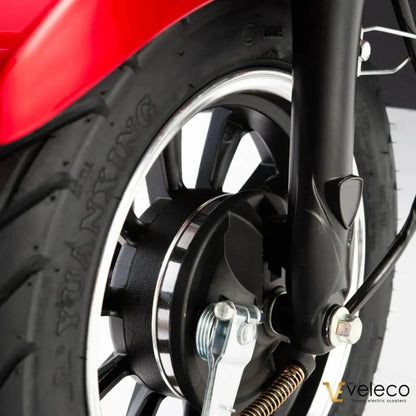 Na sliki je električni skiro znamke Veleco, model Draco. Vidna je podrobnost sprednjega kolesa, ki vključuje pnevmatiko in del sistema zaviranja. Kolo ima črno barvo s kromiranim robom, kar daje skiroju sodoben videz.