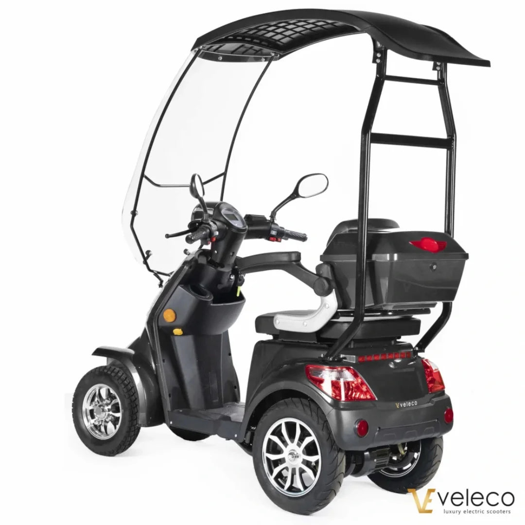 Izdelek je električni skuter, znamke Veleco, model Faster. Na sliki je vidna zasnova skuterja s tremi kolesi, zaščitnim strešnim pokrovom in udobnim sedežem. Ima tudi prostoren prostor za shranjevanje na zadnji strani ter krmilo z različnimi kontrolami. Skuter je zasnovan za udobno in varno vožnjo.