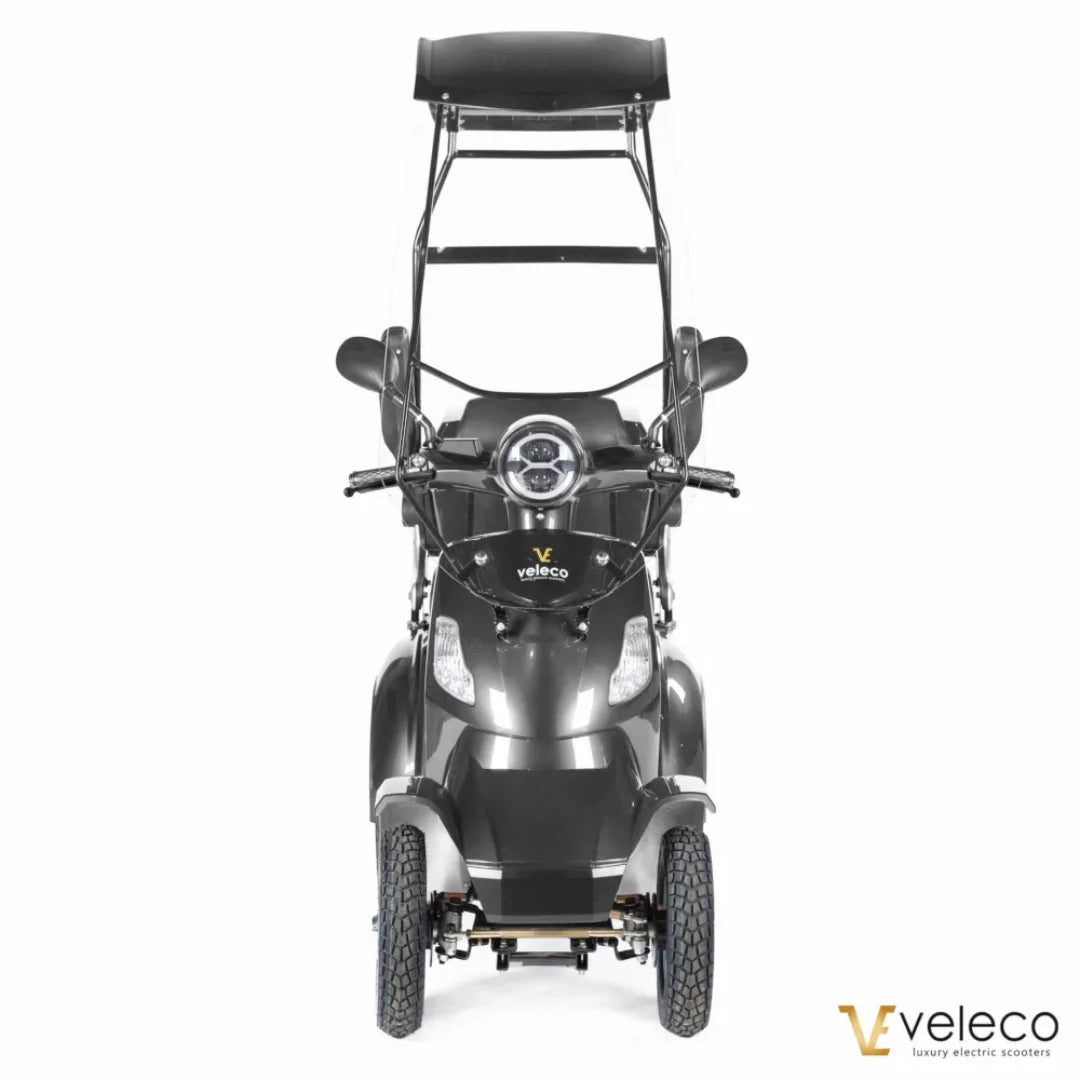 Izdelek je električni skuter, znamke Veleco, model Faster. Na sliki je prikazan sprednji del skuterja, ki ima široke ročice, okroglo luč na sredini in zaščitni strešnik nad sedežem. Skuter ima tri kolesa, kar omogoča stabilno vožnjo, in je zasnovan za udobno in enostavno uporabo.