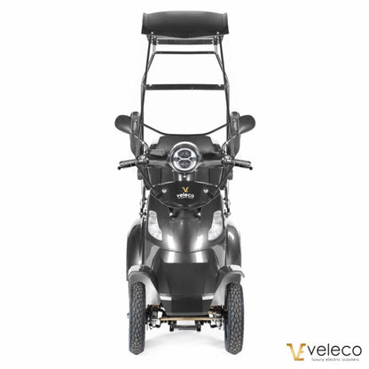 Izdelek je električni skuter, znamke Veleco, model Faster. Na sliki je prikazan sprednji del skuterja, ki ima široke ročice, okroglo luč na sredini in zaščitni strešnik nad sedežem. Skuter ima tri kolesa, kar omogoča stabilno vožnjo, in je zasnovan za udobno in enostavno uporabo.