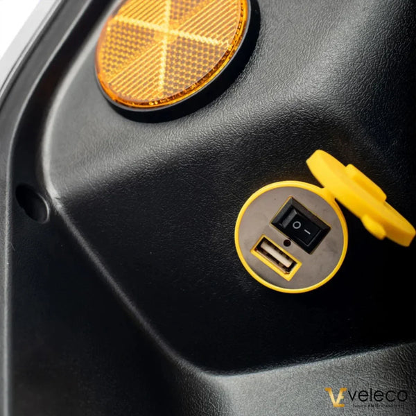 Izdelek je električni skiro znamke Veleco, model Faster. Na sliki je prikazan del skiroja, kjer se nahajajo USB priključek in stikalo, obkrožena z rumeno zaščitno kapico. Vidna je tudi odsevna oznaka v oranžni barvi, kar nakazuje na funkcionalnost in varnost izdelka.