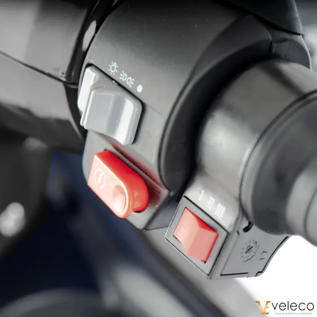 Izdelek na sliki je električni skiro, model Veleco Faster. Na sliki je prikazan del krmila, kjer so vidni različni gumbi in stikala, vključno s stikalom za luči in gumbom za vklop. Zasnova je moderna in funkcionalna, kar omogoča enostavno upravljanje.