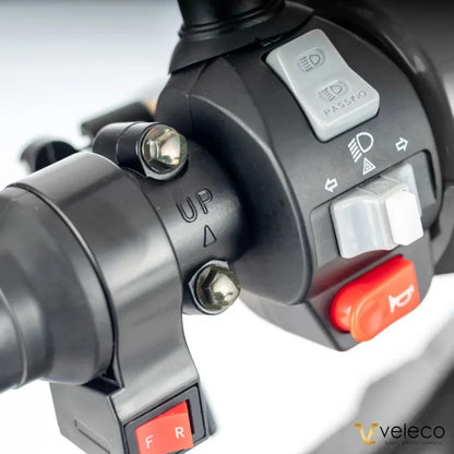 Izdelek na sliki je električni skiro znamke Veleco, model Faster. Na sliki je prikazan del krmila, kjer so vidni različni gumbi in stikala, vključno s tistimi za luči in smerokaze. Krmiljenje je zasnovano za enostavno uporabo, kar omogoča udobno vožnjo.