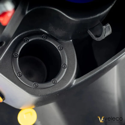 Izdelek je električni skiro znamke Veleco, model Faster. Na sliki je prikazan del skiroja, ki vključuje odprtino za polnjenje ali dostop do baterije, obkroženo z gumijastim tesnilom, kar nakazuje na vodoodpornost. Vidna je tudi rumena oznaka, ki bi lahko označevala funkcionalnost ali varnostni element.