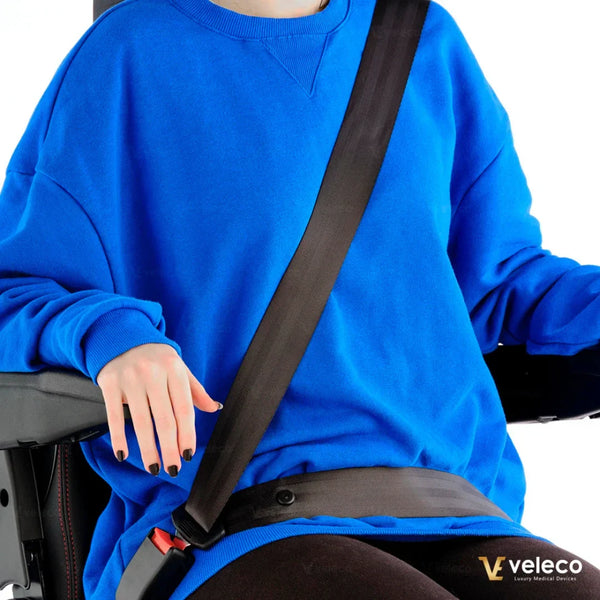 Izdelek je električni skiro, model Veleco Faster. Na sliki je oseba, ki nosi modro majico in je pripeta z varnostnim pasom na sedežu električnega skiroja. Vidna je tudi roka osebe, ki se opira na naslonjalo. Sliko zaznamuje poudarek na varnosti in udobju med vožnjo.