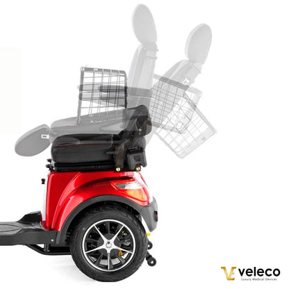 Izdelek na sliki je električni skuter, model Veleco Faster. Na sliki je vidna rdeča barva skuterja, ki ima udobno sedežno površino in naslonjalo. V ozadju je prikazana košara za shranjevanje, ki se lahko premika. Skuter je zasnovan za udobno in enostavno mobilnost.