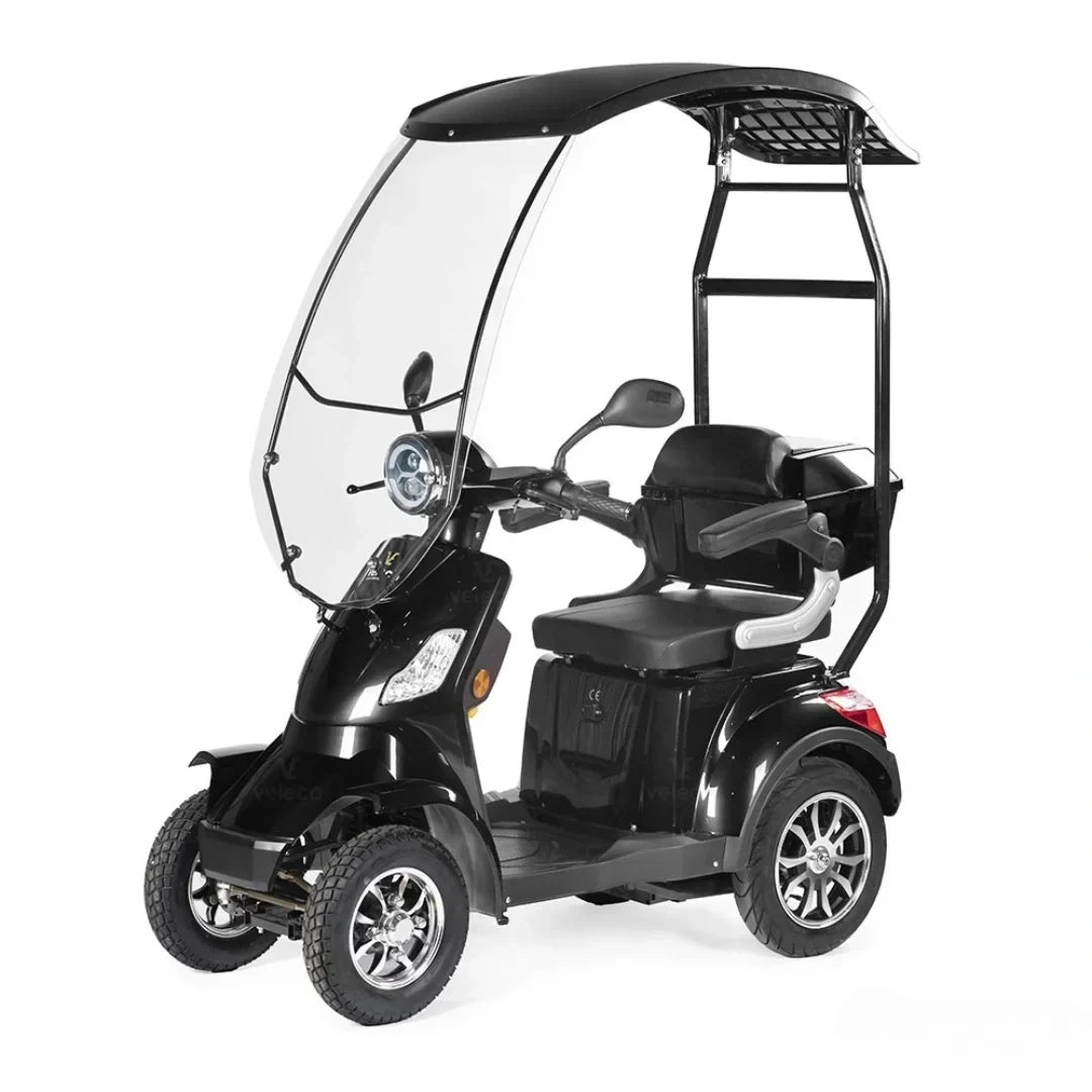 Izdelek je električni skuter, model Veleco Faster. Na sliki je prikazan črn električni skuter s tremi kolesi, ki ima zaščitno streho in vetrobransko steklo. Skuter je zasnovan za udobno vožnjo, saj vključuje široko sedežno površino in ročne naslone. Vidne so tudi kromirane platišča, kar dodaja estetski pridih.