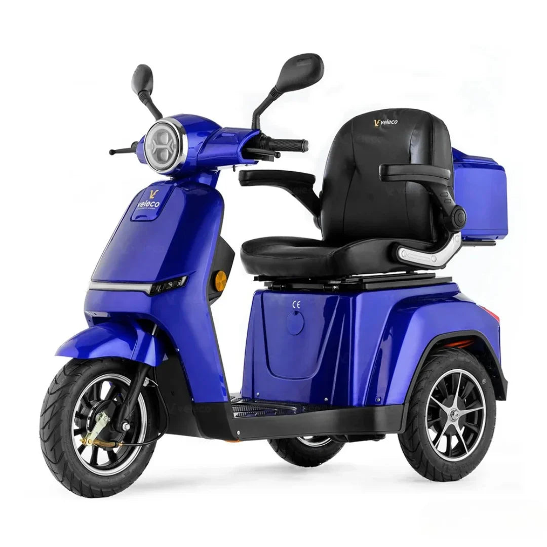 Izdelek na sliki je električni mobilni voziček, model Veleco Turris. Voziček je v svetlo modri barvi in ima udoben sedež z naslonjali za roke. Na sprednjem delu se nahajajo krmilo in luči, kar nakazuje na njegovo funkcionalnost za vožnjo. Ima tri kolesa, kar zagotavlja stabilnost, in prostor za shranjevanje.