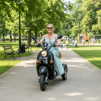 Izdelek na sliki je električni mobilni voziček, model Veleco ZT63. Na sliki je ženska, ki vozi voziček po parkovni poti, obkrožena z zelenjem in otroki, ki se igrajo v ozadju. Voziček ima črno barvo in je zasnovan za udobno vožnjo.