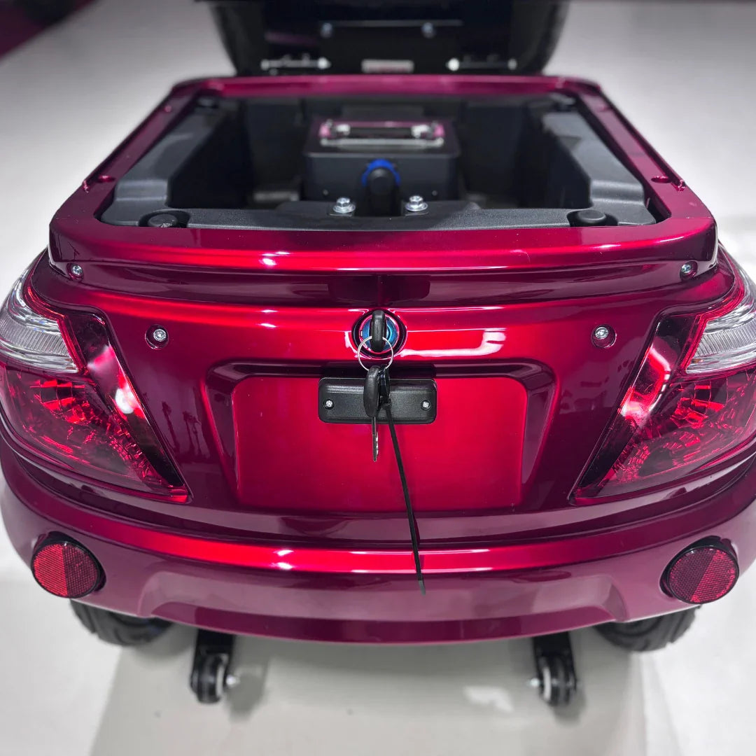 Izdelek je električni mobilni skuter na treh kolesih, model Vigorous Vista 1. Na sliki je vidna zadnja stran skuterja v svetlo rdeči barvi, kjer so prikazani zadnji luči in ključavnica. Odpiranje pokrova razkriva prostor za baterijo, kar nakazuje na funkcionalnost in zasnovo, prilagojeno za udobno vožnjo.