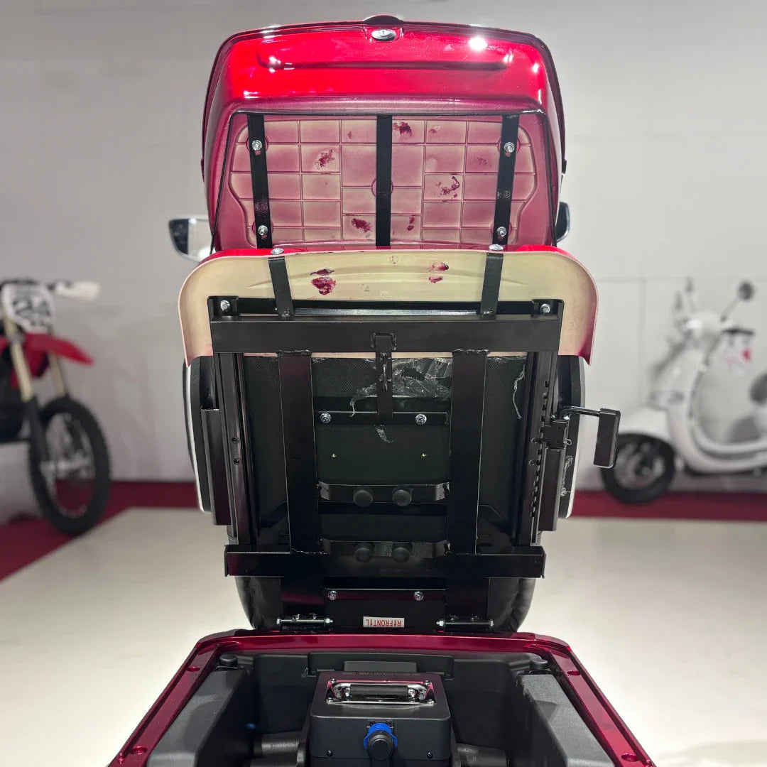 Izdelek je električni mobilni skuter s tremi kolesi, model Vigorous Vista 1. Na sliki je prikazan zadnji del skuterja, kjer je odprt pokrov, ki razkriva prostor za shranjevanje in baterijo. Skuter je v rdeči barvi, v ozadju pa so vidni tudi drugi električni skuterji.