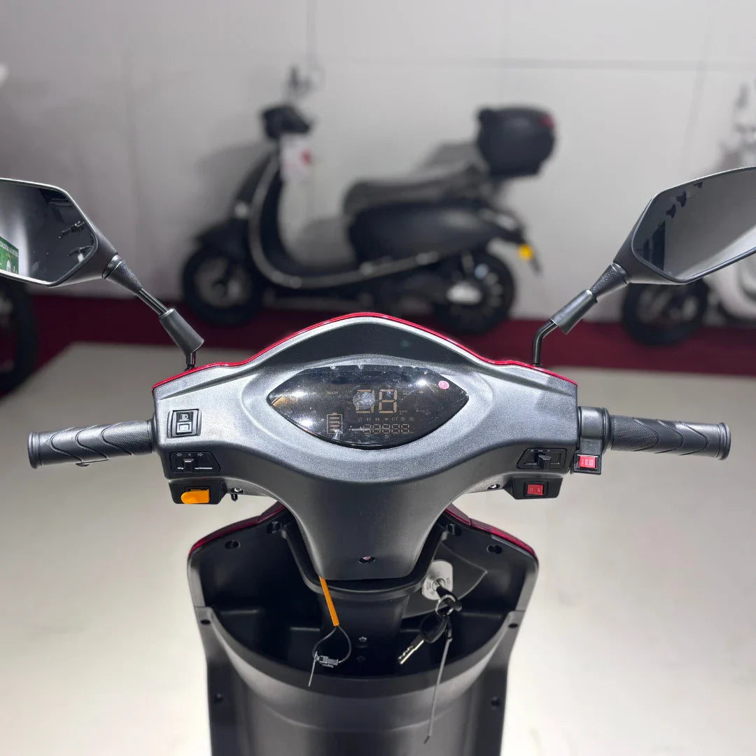 Izdelek na sliki je električni skuter z imenom Vigorous Vista 1. Na sliki je prikazan nadzorni panel, ki vključuje ročice za krmiljenje, zaslon za prikaz informacij ter ogledala. Skuter ima tri kolesa, kar omogoča stabilnejšo vožnjo, in je zasnovan za enostavno upravljanje. V ozadju so vidni tudi drugi skuterji.