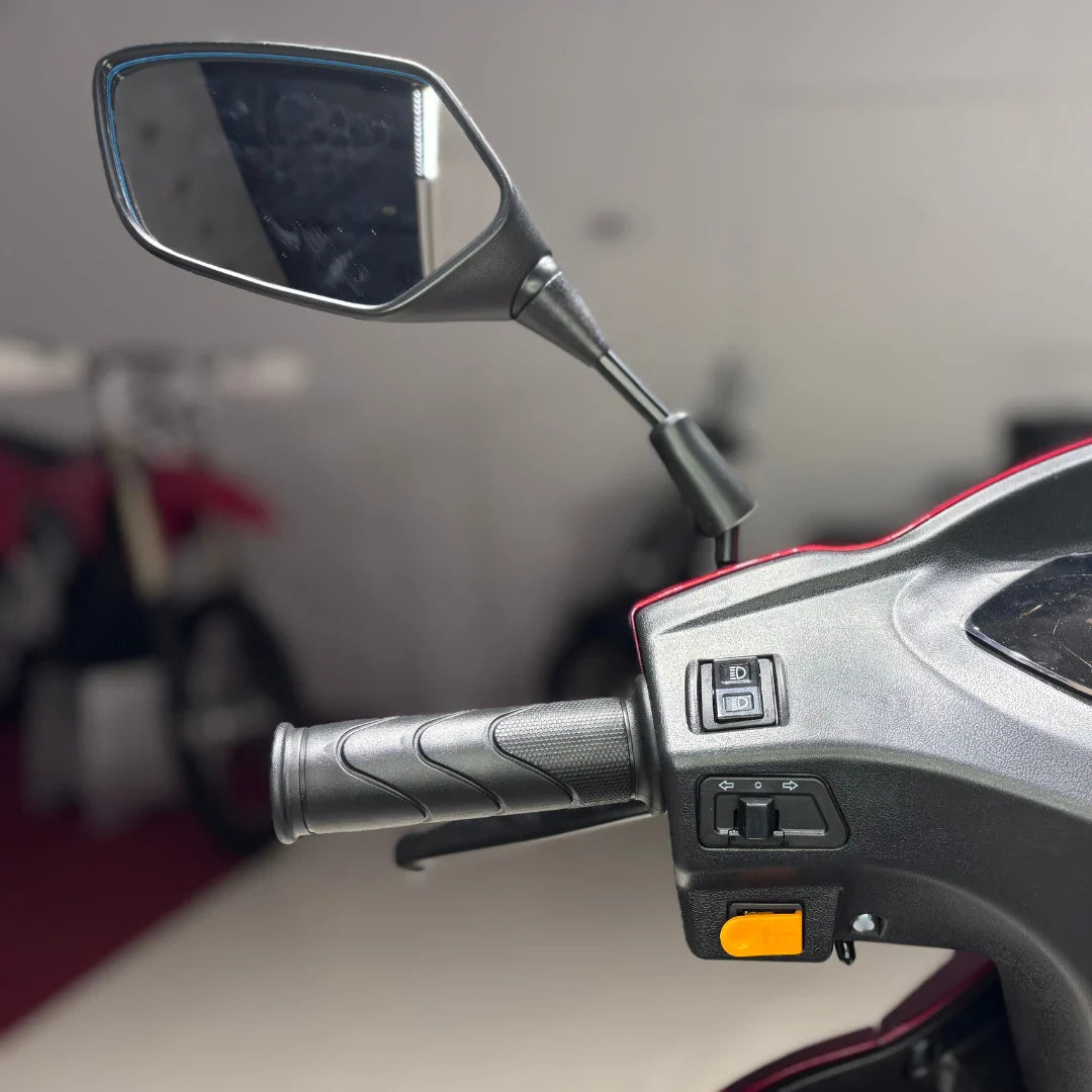 Izdelek je električni trokolni skuter, model Vigorous Vista 1. Na sliki je prikazan del krmila, kjer so vidni ročaj, ogledalo in gumbi za upravljanje. Skuter ima ergonomsko oblikovan ročaj, kar omogoča udobno vožnjo. V ozadju so vidne tudi druge motorne naprave.