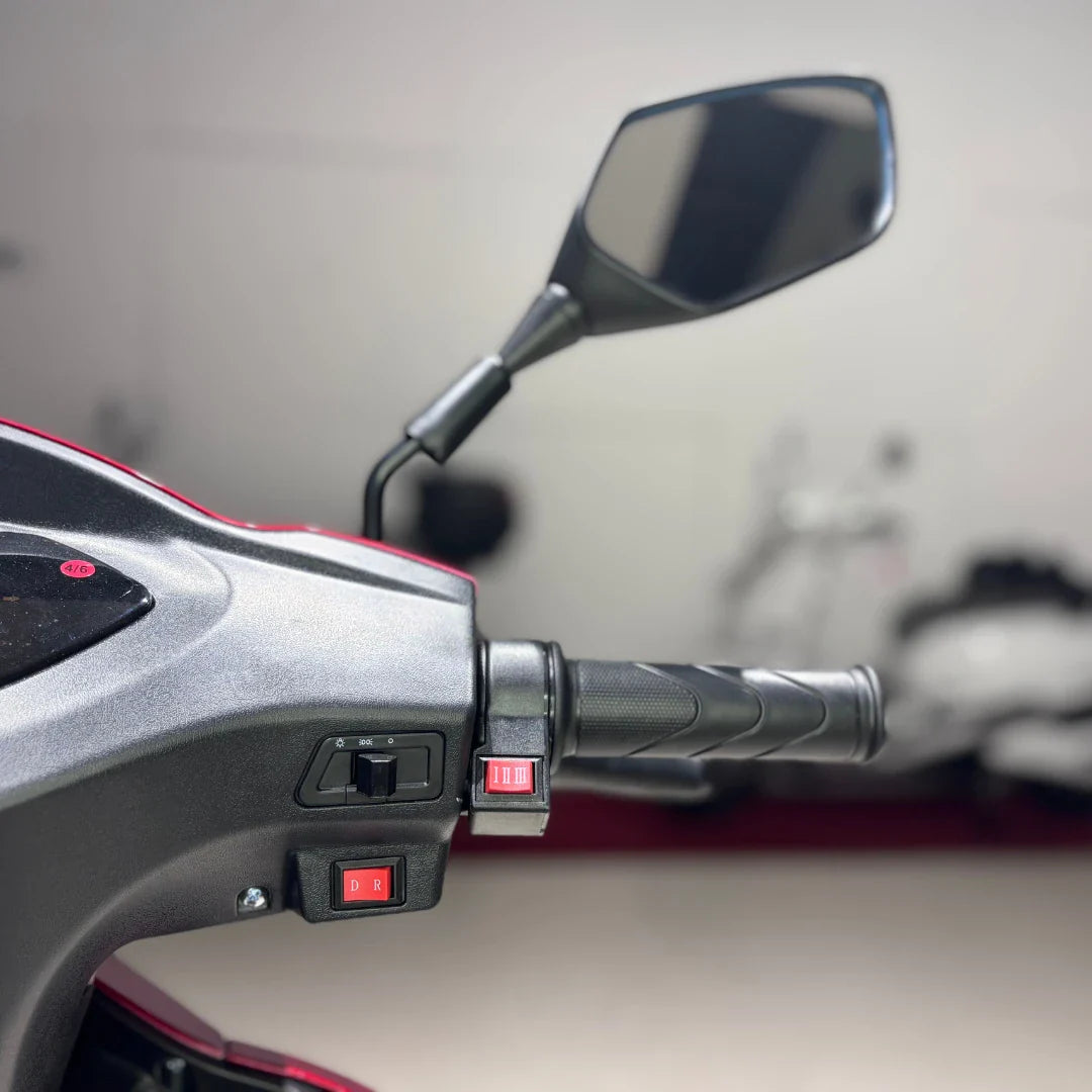 Izdelek je električni mobilni skuter s tremi kolesi, model Vigorous Vista 1. Na sliki je prikazan del krmila, kjer so vidni ročaji, stikala za prestavljanje med različnimi načini vožnje ter ogledalo. Skuter ima športen dizajn in je zasnovan za udobno vožnjo.