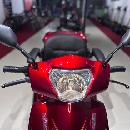 Izdelek je električni skuter na treh kolesih, model Vigorous Vista 1. Na sliki je vidna sprednja stran skuterja, ki ima svetlo in ročice za upravljanje. Barva skuterja je rdeča, kar mu daje privlačen videz. V ozadju so vidni tudi drugi električni skuterji.