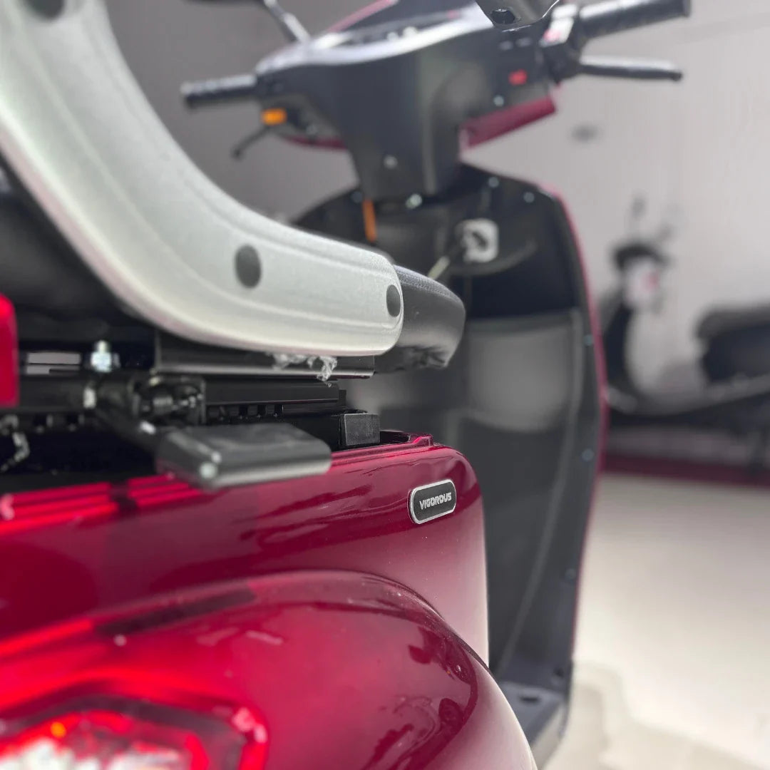 Izdelek je električni mobilni skuter s tremi kolesi, model Vigorous Vista 1. Na sliki je vidna zadnja stran skuterja v rdeči barvi, s sedežem in delom za nadzor. Dodatno je prikazana oznaka znamke "Vigorous" na telesu skuterja. V ozadju se vidi še del drugega skuterja.