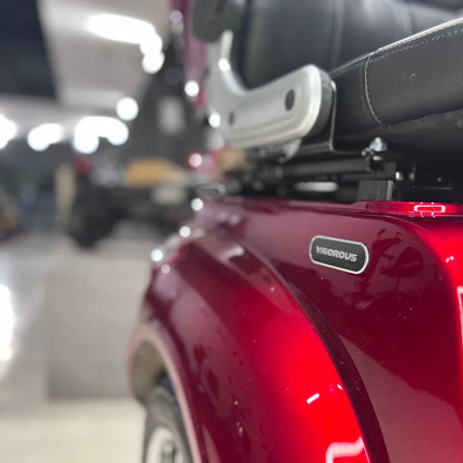 Izdelek na sliki je električni mobilni skuter s tremi kolesi, model Vigorous Vista 1. Na sliki je vidna rdeča karoserija skuterja, ki ima na stranski strani oznako znamke "Vigorous". V ozadju se vidi del opreme, ki nakazuje, da je skuter postavljen v notranjem prostoru, verjetno v trgovini ali razstavnem prostoru.