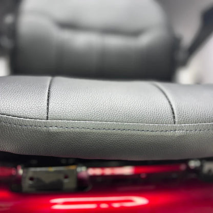 Izdelek je električni mobilni skuter, model Vigorous Vista 1. Na sliki je prikazan sedež skuterja, ki je oblečen v sivo umetno usnje. Vidna je tekstura sedeža, ki je udoben in zasnovan za dolgotrajno uporabo. V ozadju se nahaja del skuterja, ki je rdeče barve.