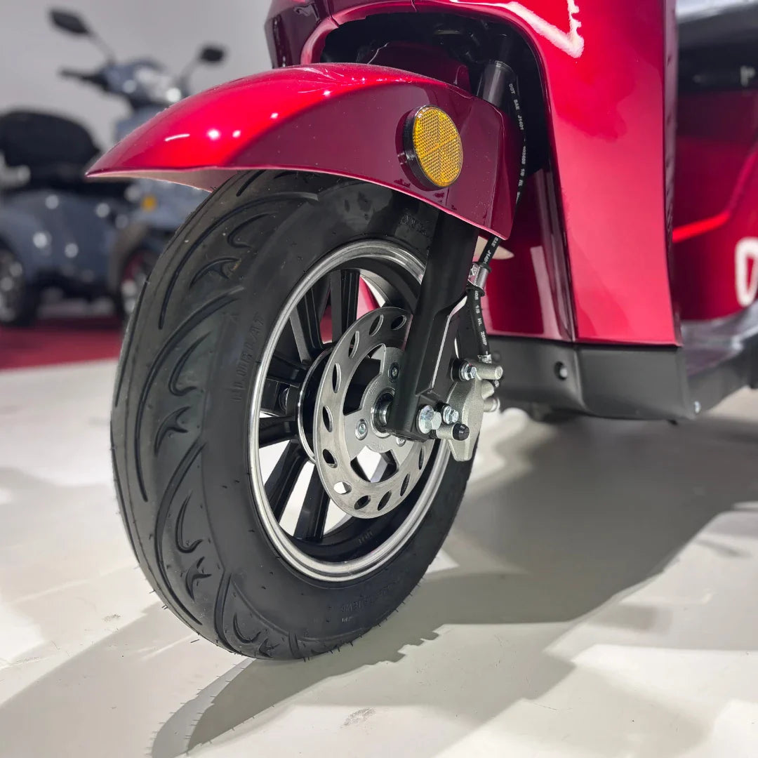 Izdelek na sliki je električni mobilni skuter s tremi kolesi, model Vigorous Vista 1. Na sliki je prikazan sprednji del skuterja, ki vključuje rdečo karoserijo, sprednje kolo ter disk zavore. Kolo ima široko pnevmatiko in je zasnovano za stabilno vožnjo. V ozadju so vidni tudi drugi skuterji, kar nakazuje, da gre za razstavni prostor.