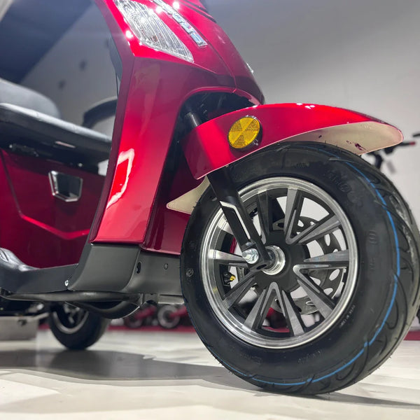 Izdelek je električni skuter s tremi kolesi, model Vigorous Vista 1. Na sliki je vidna sprednja kolesna enota, ki vključuje črno pnevmatiko z modrim obodom ter svetlobno oznako na sprednjem delu. Skuter je v svetlo rdeči barvi, kar mu daje privlačen videz. V ozadju je del sedeža in notranjosti skuterja, kar nakazuje na udobno zasnovo.