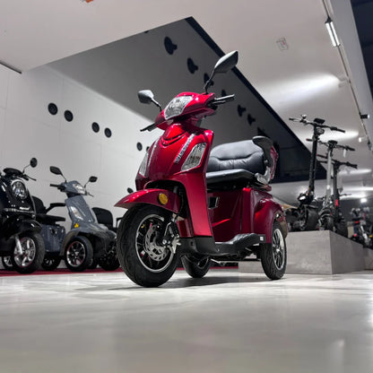 Izdelek na sliki je električni mobilni skuter, model Vigorous Vista 1. Skuter ima tri kolesa in je v rdeči barvi, kar mu daje dinamičen videz. Na sliki je jasno vidna sprednja stran skuterja z udobnim sedežem in krmilom, v ozadju pa so vidni tudi drugi podobni električni skuterji. Celotna scena se odvija v sodobnem prostoru, kar dodatno poudarja modernost izdelka.