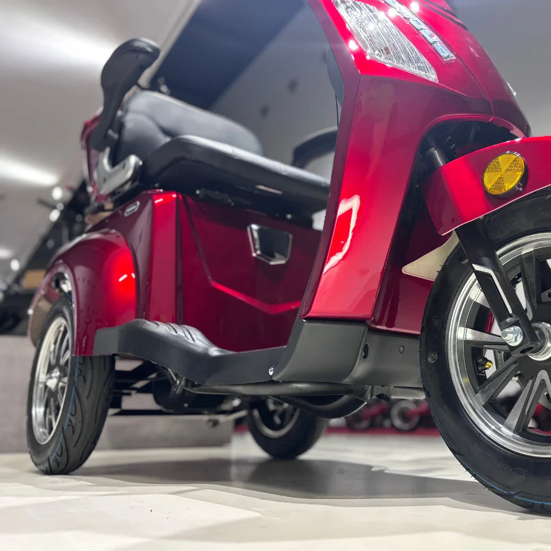 Izdelek je električni skuter s tremi kolesi, model Vigorous Vista 1. Na sliki je vidna rdeča karoserija skuterja, udobno sedežno mesto ter sprednje kolo z značilnim dizajnom. Osvetlitev in detajli na karoseriji dajejo sodoben videz, medtem ko je ozadje nevtralno, kar poudarja izdelek.