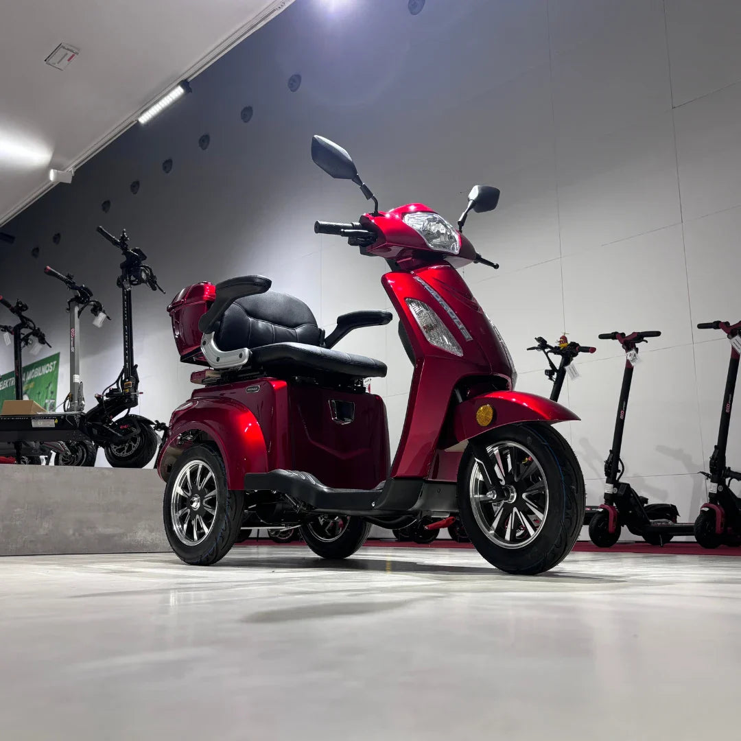 Izdelek na sliki je električni skuter s tremi kolesi, model Vigorous Vista 1. Skuter je v svetlo rdeči barvi in ima udobno sedežno površino s hrbtno oporo. Na zadnjem delu je nameščen prtljažnik, kar omogoča dodatno shranjevanje. V ozadju so vidni tudi drugi električni skuterji, kar nakazuje, da gre za prodajno mesto ali razstavo.