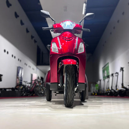 Izdelek na sliki je električni skuter s tremi kolesi, model Vigorous Vista 1. Na sliki je jasno viden sprednji del skuterja, ki je v svetlo rdeči barvi, z izrazitimi lučmi in krmilom. V ozadju so vidni tudi drugi električni skuterji in kolesa, kar nakazuje, da je slika posneta v prodajnem prostoru.