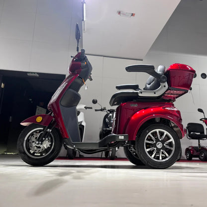 Izdelek na sliki je električni mobilni skuter s tremi kolesi, model Vigorous Vista 1. Ima rdečo karoserijo in udobno sedežno površino z naslonjali. Na sprednjem delu so vidni krmilo in ogledalo, medtem ko je na zadnjem delu skuterja prtljažnik. Skuter je zasnovan za enostavno in udobno vožnjo, kar ga dela primernega za različne uporabnike.