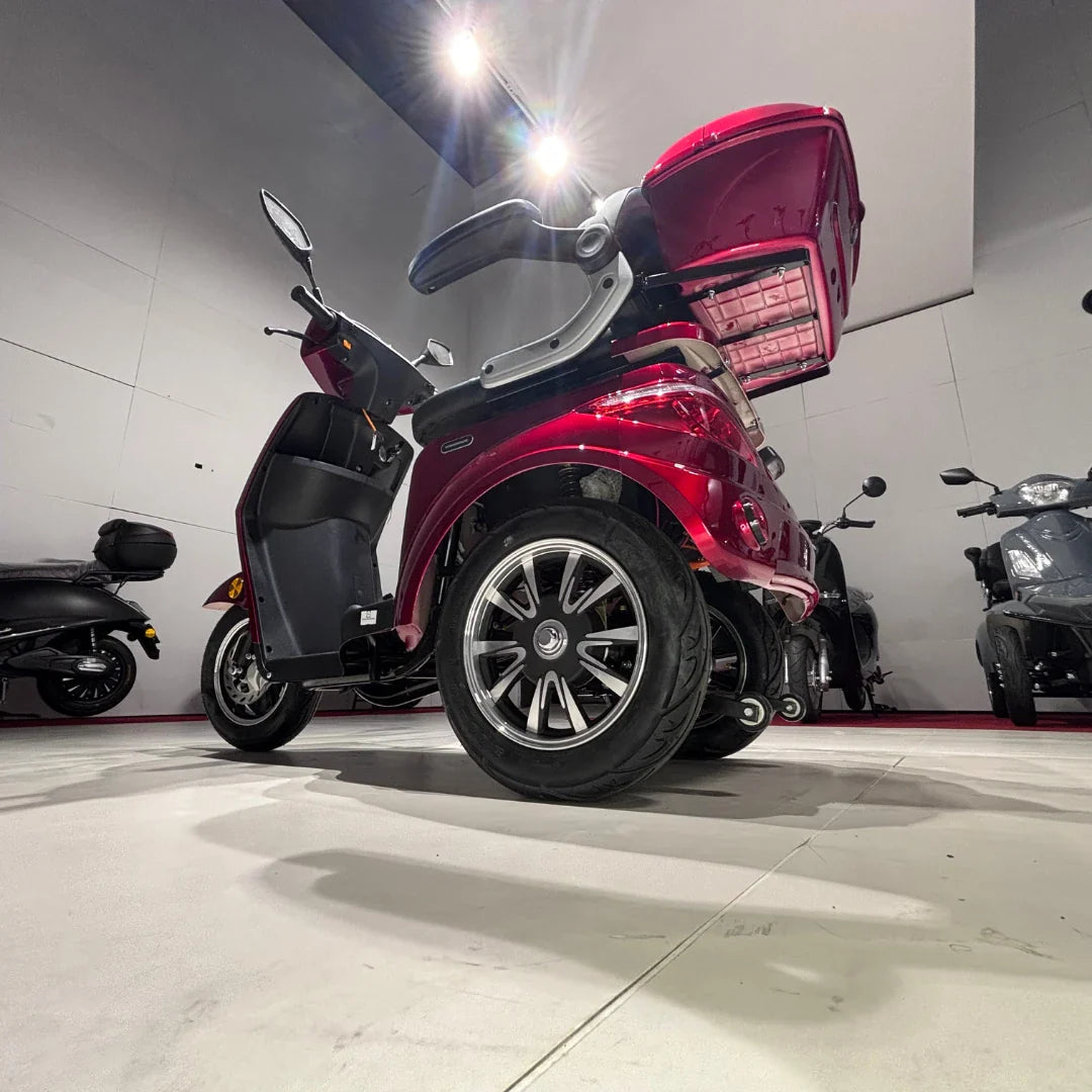 Na sliki je prikazan trikolni električni mobilnostni skuter, model Vigorous Vista 1. Skuter je v rdeči barvi in ima udobno sedežno mesto ter prostoren prtljažnik, ki je odprt. Vidni so tudi veliki kolesi s sodobnim dizajnom. V ozadju so še drugi podobni skuterji, kar nakazuje, da gre za razstavo ali prodajno mesto.