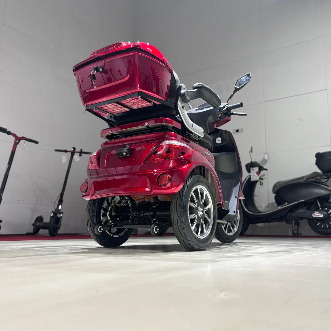 Izdelek na sliki je električni mobilni skuter s tremi kolesi, model Vigorous Vista 1. Skuter ima svetlo rdečo barvo in je zasnovan za udobno vožnjo. Vidna je tudi prtljažna enota na zadnjem delu, kar omogoča shranjevanje osebnih predmetov. V ozadju so vidni tudi drugi električni skuterji, kar nakazuje na okolje, kjer se ti izdelki prodajajo ali razstavljajo.