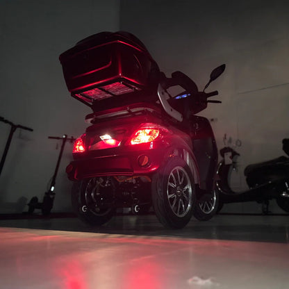 Izdelek na sliki je električni skuter s tremi kolesi, model Vigorous Vista 1. Na sliki je prikazan zadnji del skuterja, ki ima rdeče luči in prostoren prtljažnik na vrhu. Opremljen je z udobnim sedežem in dodatnimi funkcijami, ki olajšajo vožnjo. V ozadju so vidni tudi drugi električni skuterji.